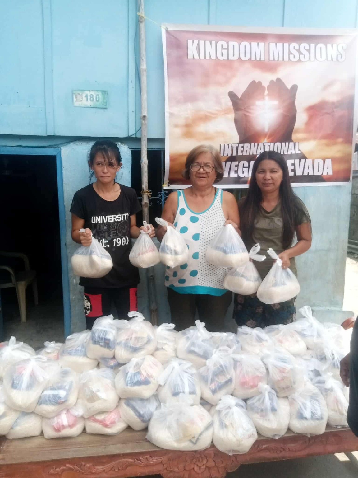 Three women holding packages of food beneath a Kingdom Missions International banner during a community food distribution